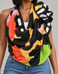 A multicolor abstract print scarf with black, orange, yellow, green, and white patterns, styled as a cowl neck drape on a model wearing blue jeans.