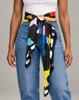 A person wearing blue jeans and a white top, shown from the chest down, with a colorful abstract-patterned silk scarf tied around the waist like a belt. The scarf features black, yellow, blue, pink, orange, and white shapes.