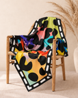 A square scarf with a vibrant rainbow watercolor splatter background and black leopard spots, finished with a white dashed border, shown on chair