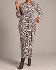 A woman modeling a long-sleeve maxi dress with a dark brown and white zebra print, featuring a self-tie waist belt and side pockets, paired with dark brown heeled boots against a neutral gray studio background.