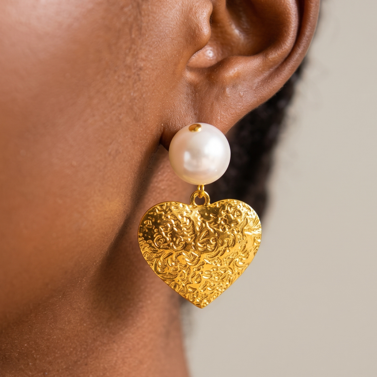 Close up of Amora Heart Dangle Earring showcasing an oversized round pearl above a textured gold colored heart pendant.