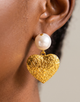 Close up of Amora Heart Dangle Earring showcasing an oversized round pearl above a textured gold colored heart pendant.