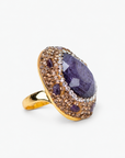 Purple statement ring featuring a faceted stone surrounded by crystals and textured Gold tone finish details.