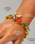 Gold colored stretch bracelet featuring sculptural hummingbird motifs, green enamel leaves and a coral flower centerpiece.