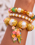 Three piece tropical bracelet stack featuring luminous pearls, colorful artisan beads, floral accents, butterfly charms, and a detailed tropical parrot charm in gold tone.