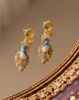 Statement peacock earrings featuring hand painted enamel feathers, sparkling crystal accents, gold shell studs, and colorful jewel toned detailing.