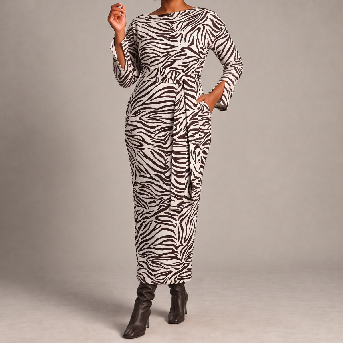 A woman modeling a long-sleeve maxi dress with a dark brown and white zebra print, featuring a self-tie waist belt and side pockets, paired with dark brown heeled boots against a neutral gray studio background.