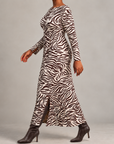 A woman modeling a long-sleeve maxi dress with a brown and white zebra print, paired with dark brown heeled ankle boots, against a neutral gray studio background.