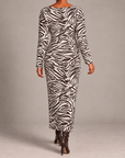 A woman modeling a long-sleeve, bodycon maxi dress with a dark brown and white zebra print, paired with dark brown heeled boots, shot from behind against a neutral gray studio background.