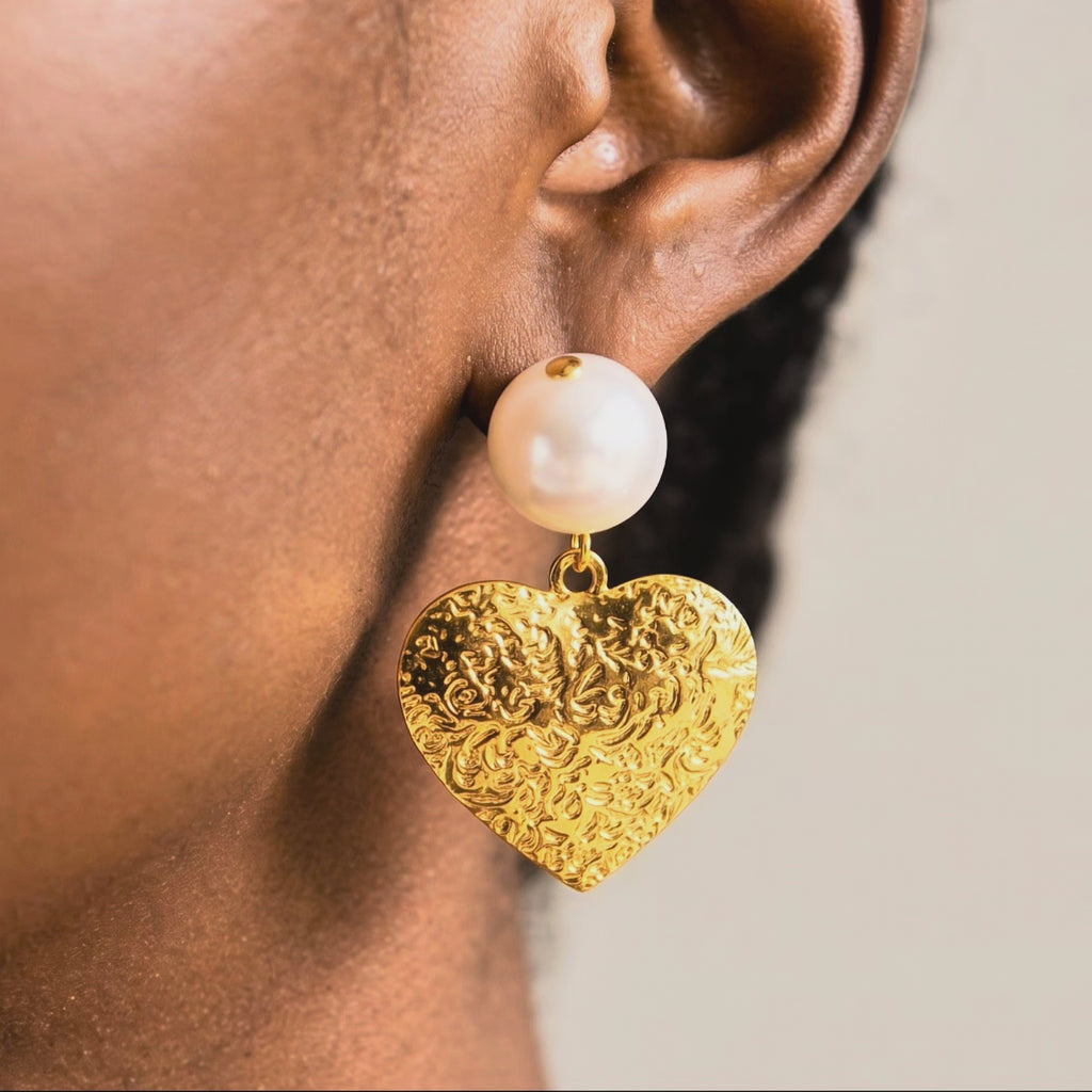 Romantic statement dangle earrings featuring ivory pearls and polished gold hearts with raised decorative texture.