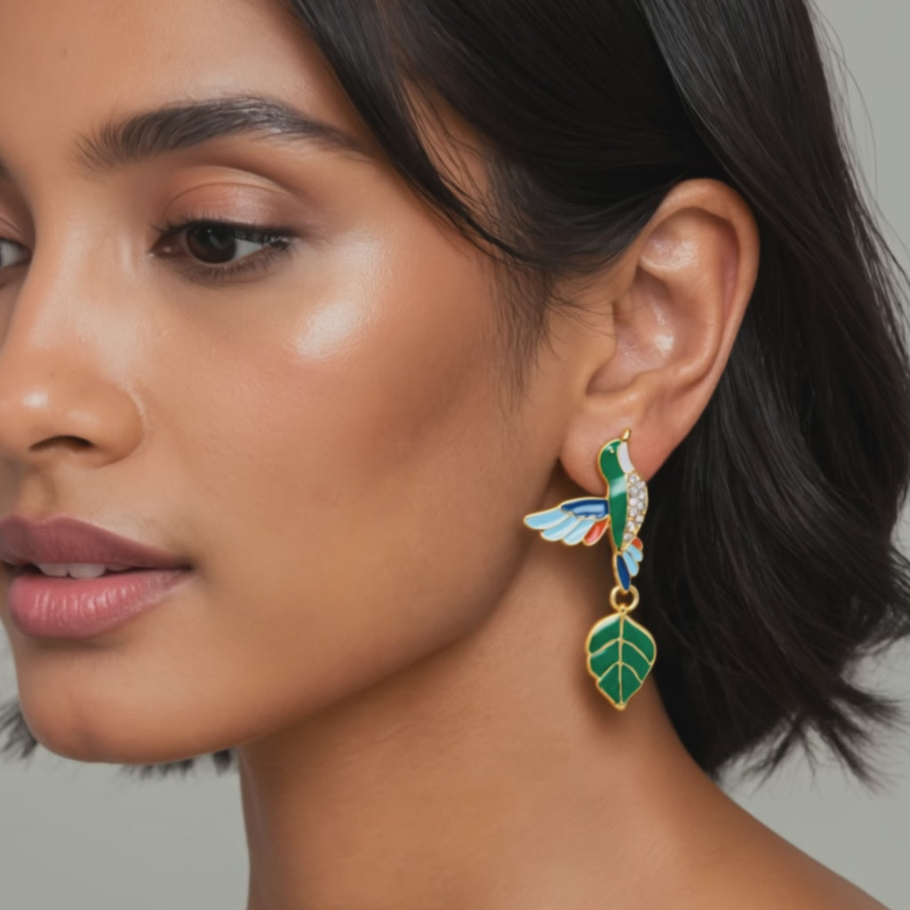 Gold colored statement dangle earrings featuring hummingbird motifs, glossy green enamel leaves and colorful wings.