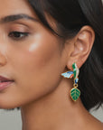 Gold colored statement dangle earrings featuring hummingbird motifs, glossy green enamel leaves and colorful wings.
