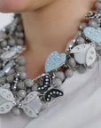 Sassy Jones Lovebug Greige Bib featuring whimsical dimensional motifs in powder blue, soft gray, black and neutral tones.